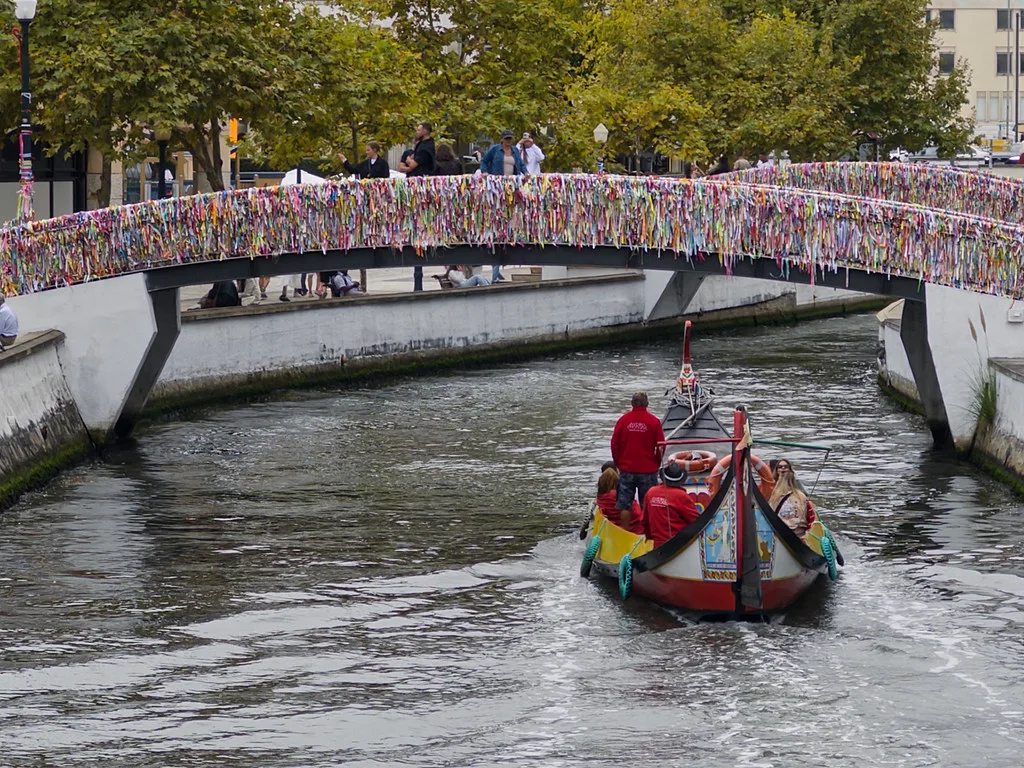 A moliceiro boat passing under a ribbon-decked bridge on a canal in Aveiro.