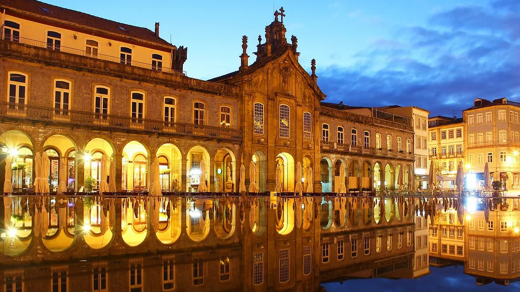 Arcaded buildings in central Braga lit at night and reflected in the water.