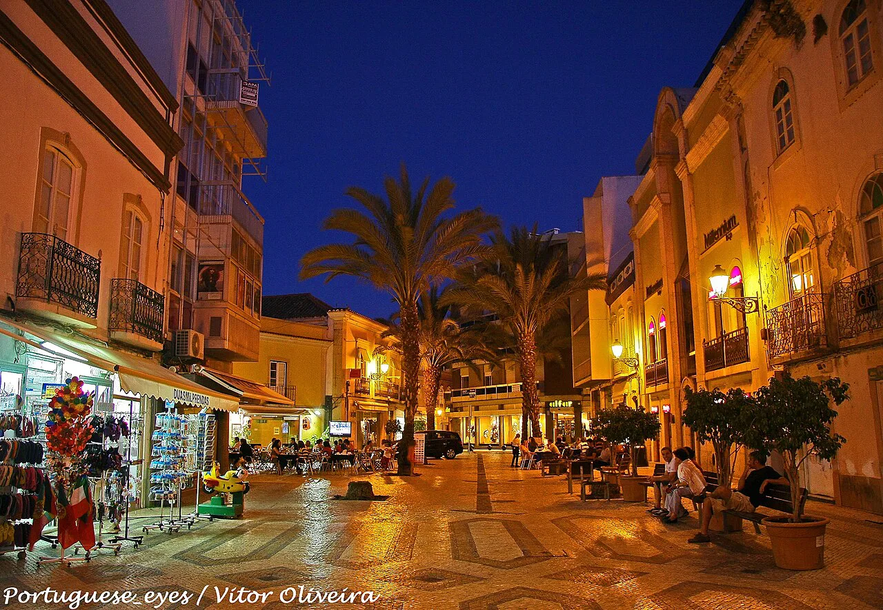 A paved street in Faro at night, lined with shops and palm trees.