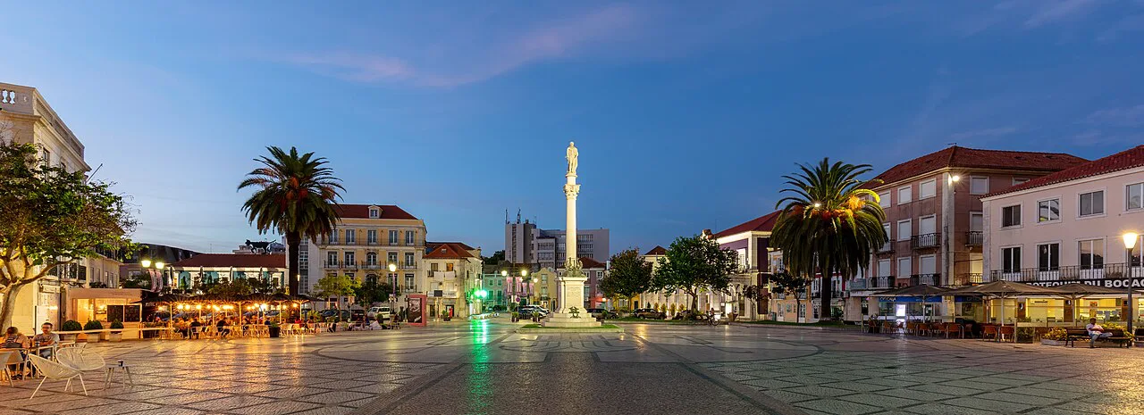 Praça do Bocage in Setúbal at dusk, with palm trees and a column.
