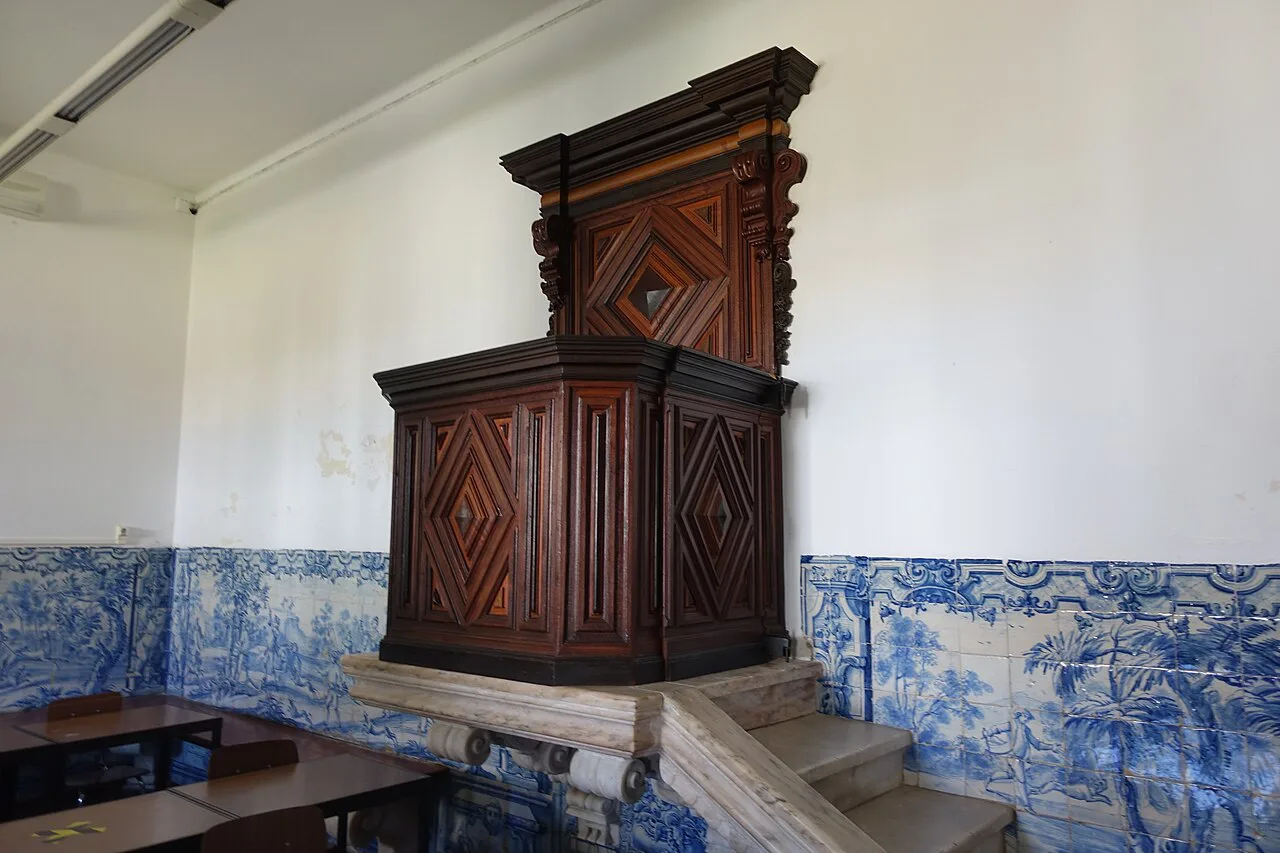 A carved wooden pulpit above blue-and-white azulejo panels in a classroom at the University of Évora.